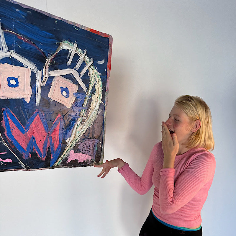 Woman (Clara Woods) in a pink sweater standing next to a large abstract painting on a white wall. Artwork Awkwardness by Clara Woods