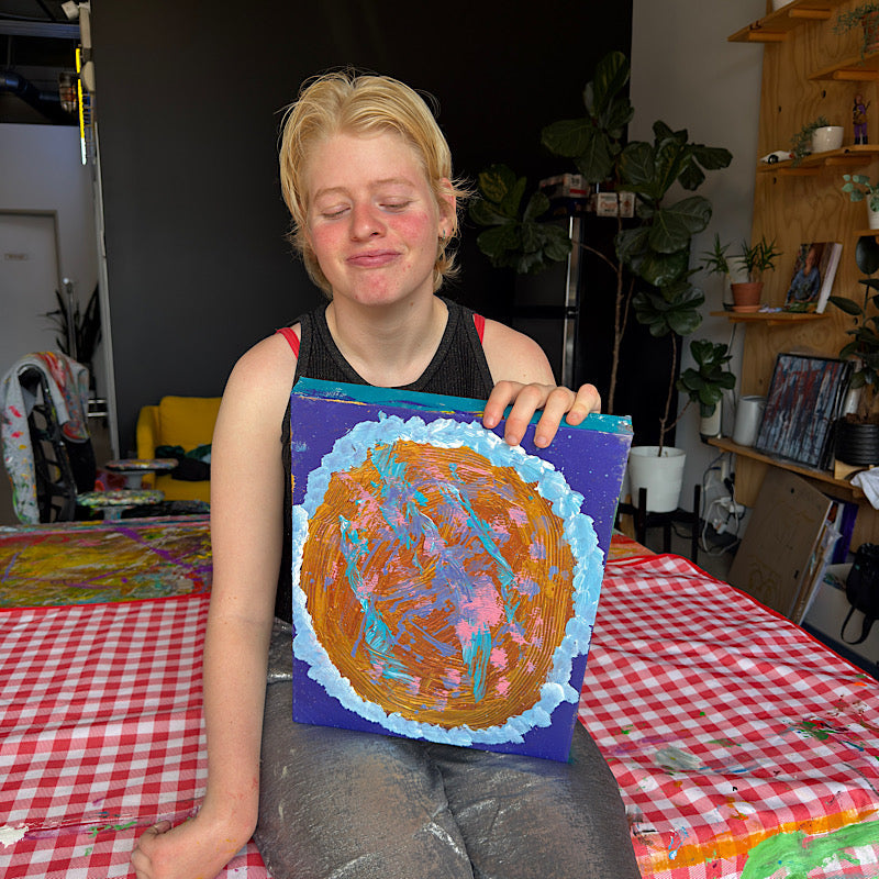 Clara Woods sitting in her studio proudly holding Little Jupiter original painting, gold and teal planet on deep navy canvas, 16x12 in, 2026