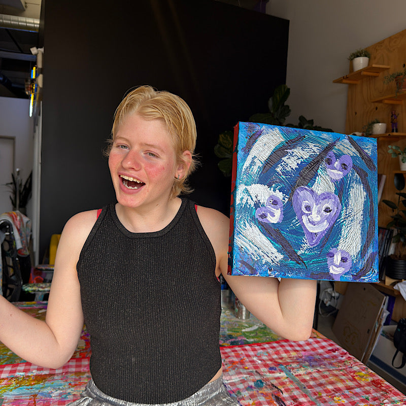 Clara Woods laughing with pure joy holding Little Neptune original painting in her studio, deep blue and teal canvas with smiling purple heart motifs, 12x12 in, 2026