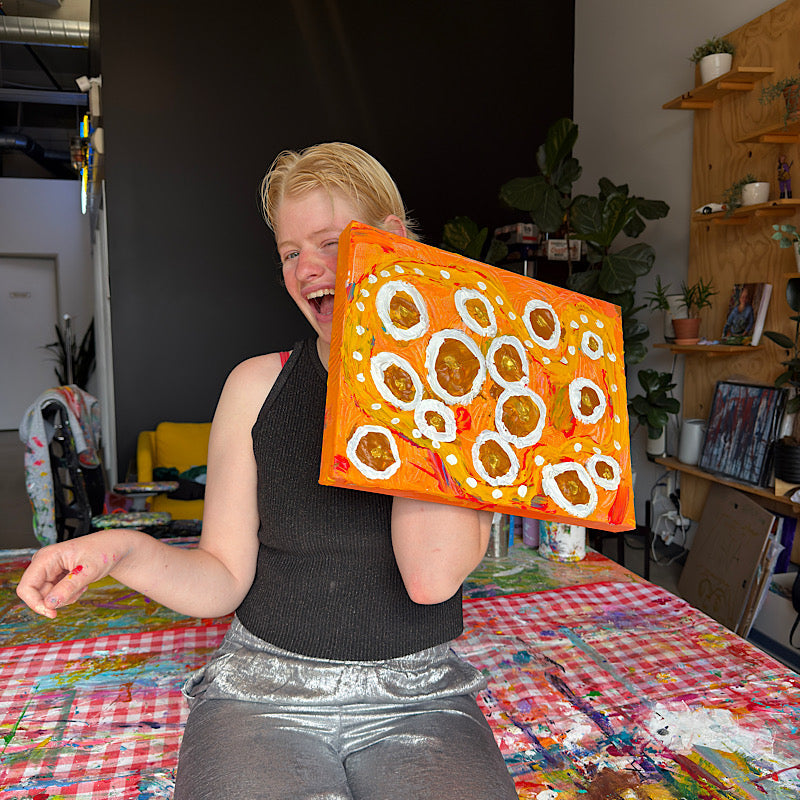 Clara Woods joyfully posing with Little Saturn original painting in her studio, vibrant orange and gold canvas with white ring motifs, 16x12 in, 2026