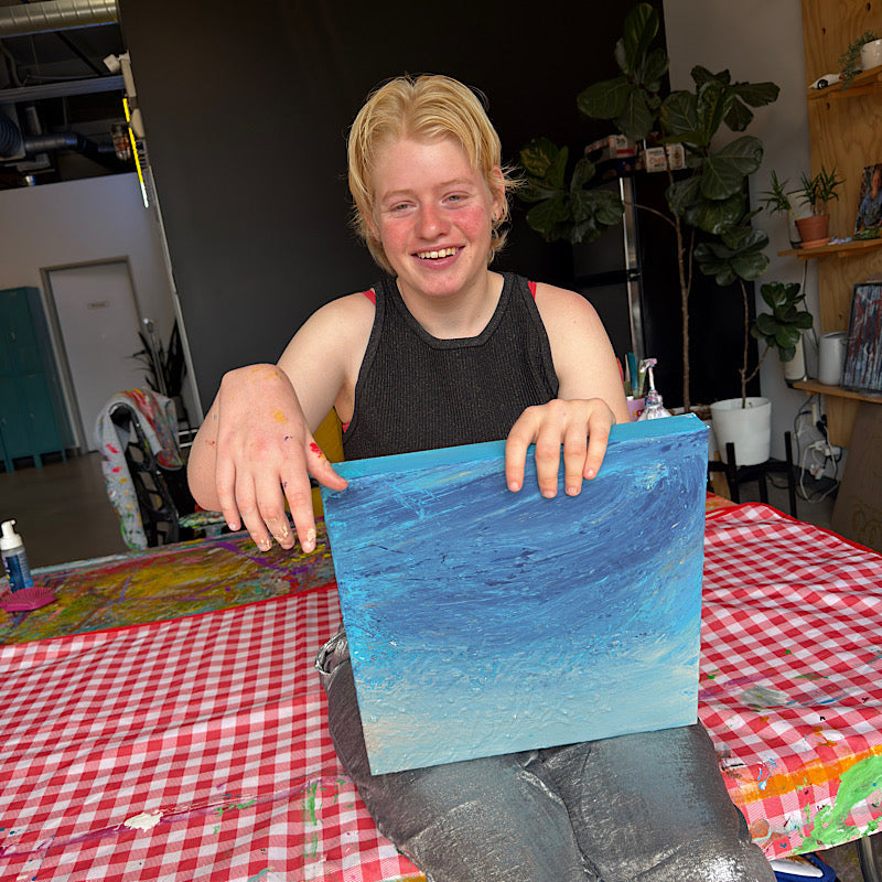 Clara Woods smiling and holding Little Uranus original painting in her studio, icy blue and teal canvas, 14x14 in, 2026