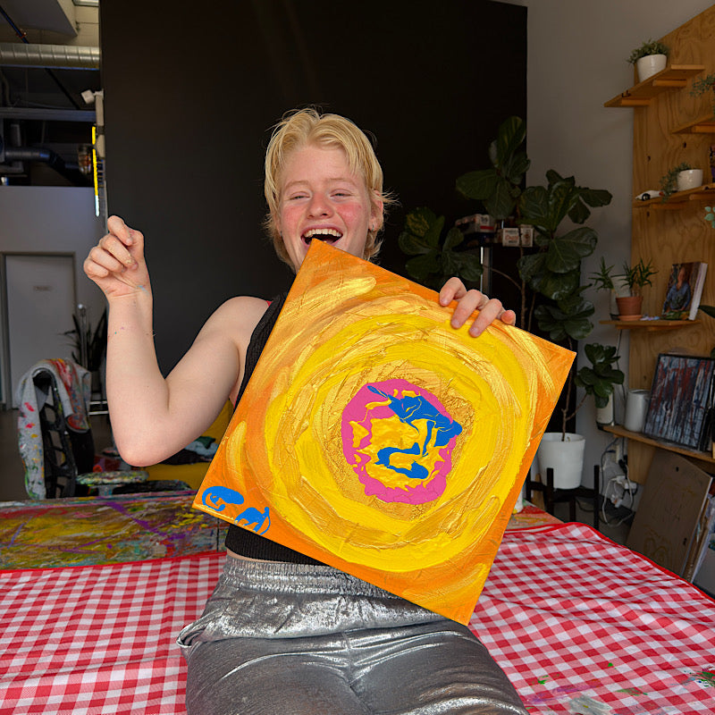Clara Woods laughing and holding Little Venus original painting in her studio, warm yellow and gold swirling canvas with pink and blue center, 14x14 in, 2026