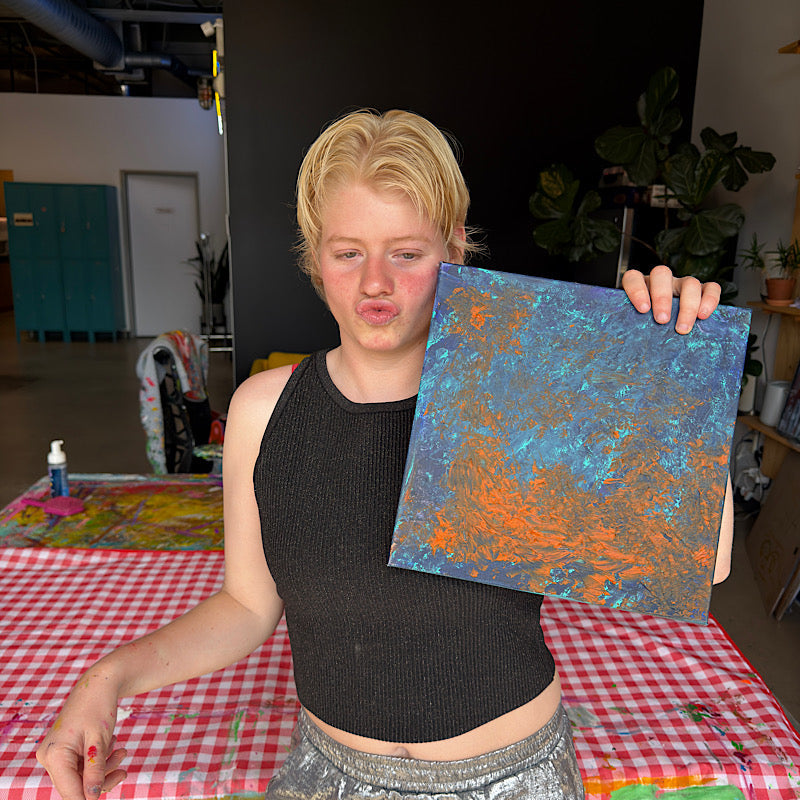 Clara Woods holding Little Mercury original painting in her studio, deep blue and orange abstract planet, 12x12 in, 2026