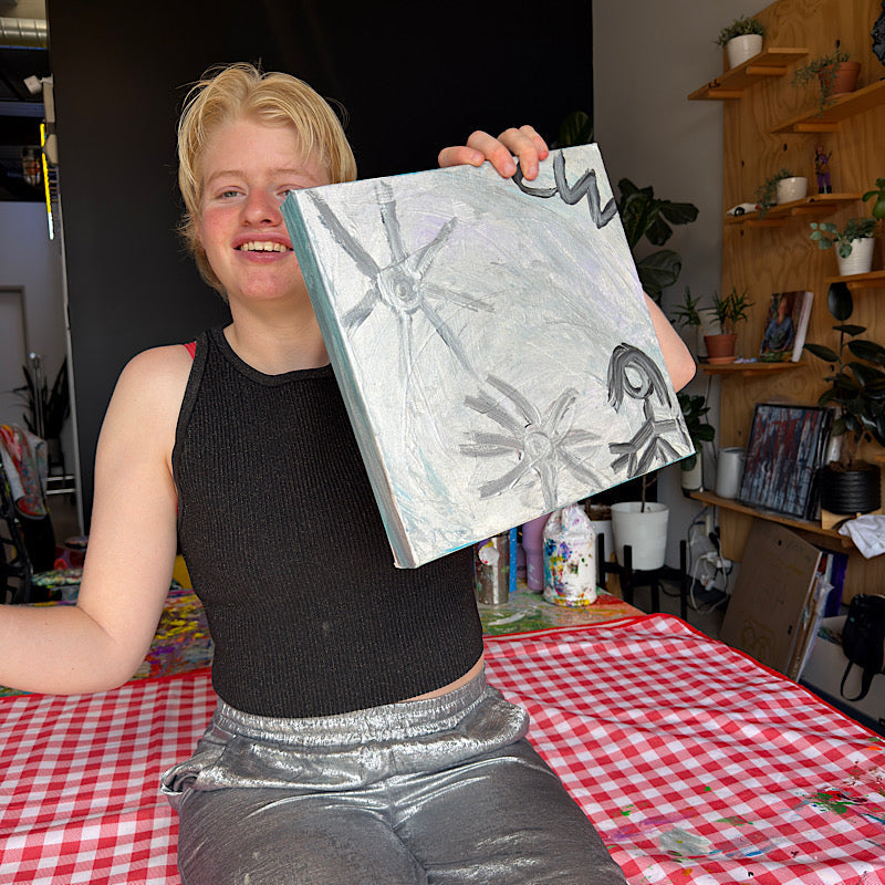 Clara Woods smiling and holding Little Moon original painting in her studio, silver and lavender canvas with star motifs, 12x12 in, 2026