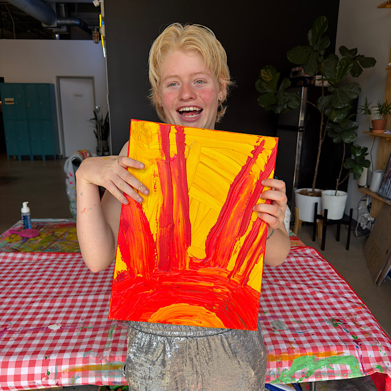 Clara Woods laughing and holding Little Sun original painting in her studio, fiery yellow and red acrylic sun with bold rays, 16x12 in, 2026